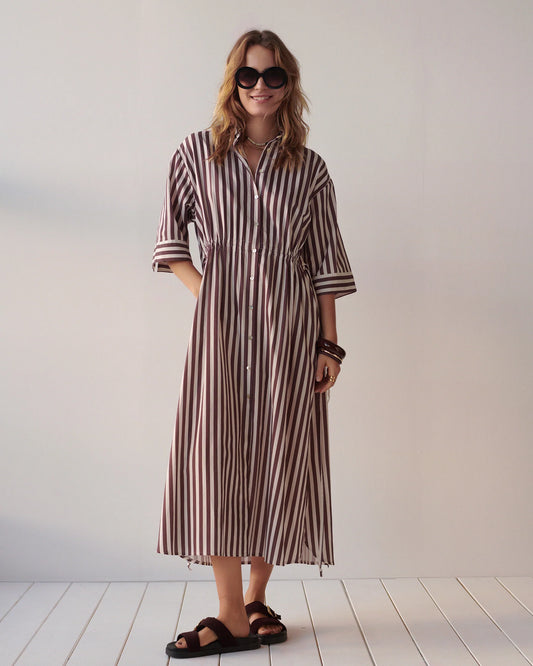 A woman modeling a brown and white vertically striped midi shirt dress with three-quarter sleeves, button-front closure, and a relaxed fit, styled with dark sunglasses, layered necklaces, bracelets, and dark sandals against a plain light wall and white floorboards.